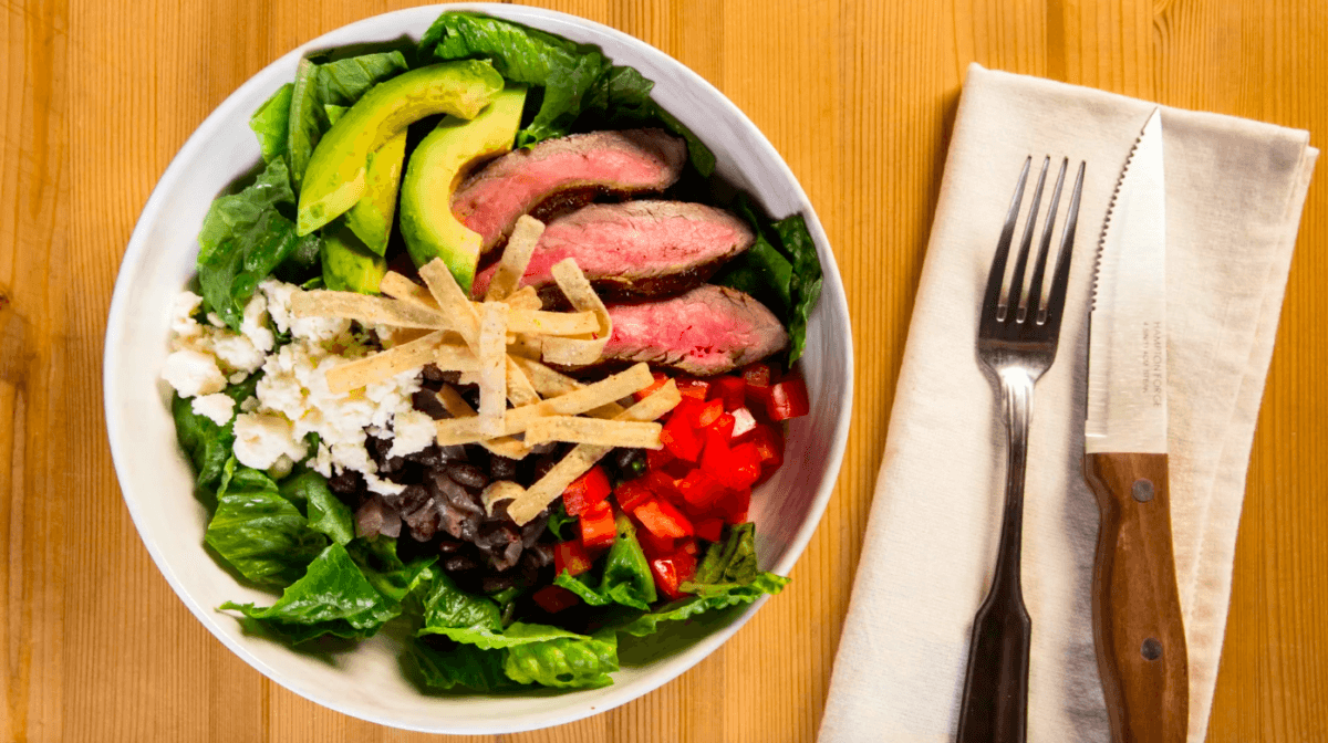 Carne asada salad bowl next to napkin with fork & knife.